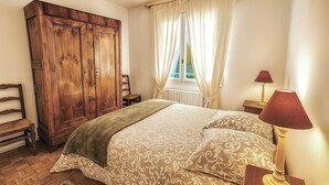 Una camera da letto con un armadio in legno, un letto con biancheria decorata, una sedia, una finestra con tende e una lampada da comodino.