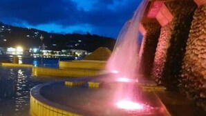 Une vue nocturne d’un élément décoratif aquatique agrémenté d’une fontaine, entouré de piliers en pierre et illuminé par des lumières roses.