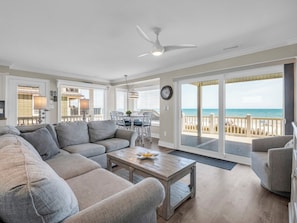 A living room with a large gray sectional sofa, a wooden coffee table, and a view of the ocean through a sliding glass door.