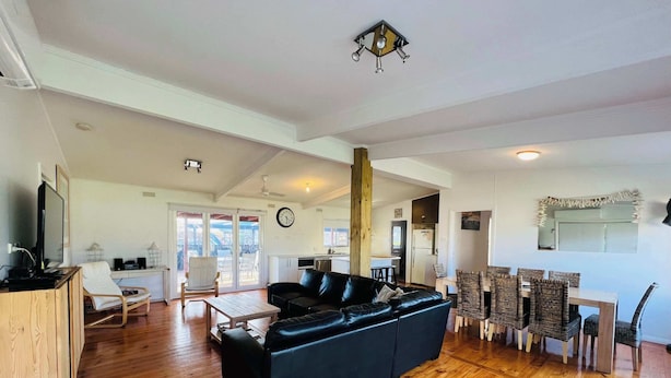 A spacious living area with a black leather sofa, wooden flooring, a dining table with wicker chairs, and a kitchen in the background.