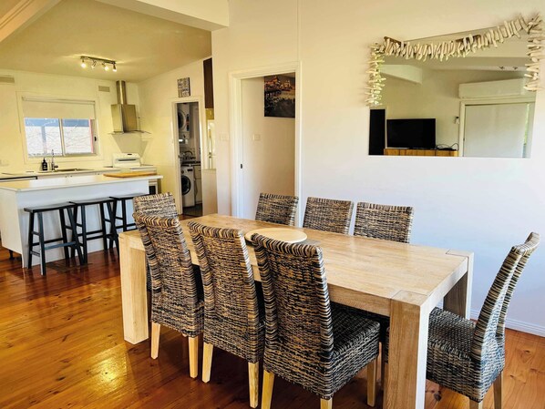 A dining area with a wooden table and wicker chairs, a kitchen in the background, and a large mirror on the wall. - Teddy's Shack  -Pet friendly outside (Robe)