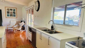 A kitchen with white cabinets, a stainless steel sink, a dishwasher, and a stove. There is a clock on the wall and a window with a view of the outside.