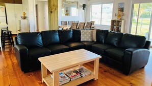A modern living room with a black leather sectional sofa, a wooden coffee table with magazines, a dining area with wicker chairs, and large windows offering a view.