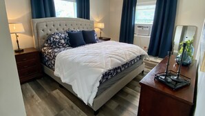 Een slaapkamer met een gestoffeerd hoofdeinde, een groot bed met een witte deken, twee donkerblauwe kussens, een houten nachtkastje met een lamp, een plafondventilator en een raam met blauwe gordijnen.