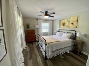 Une chambre à coucher avec un lit, une commode, un ventilateur de plafond, une fenêtre avec des rideaux et un tableau au mur.