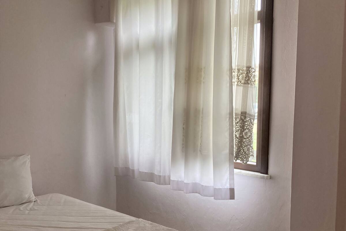 A bedroom with a bed, a window with curtains, and a wall-mounted thermostat.