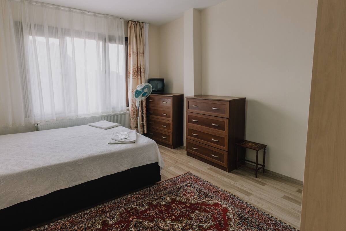 A bedroom with a bed, a dresser, a small table, and a window with curtains.