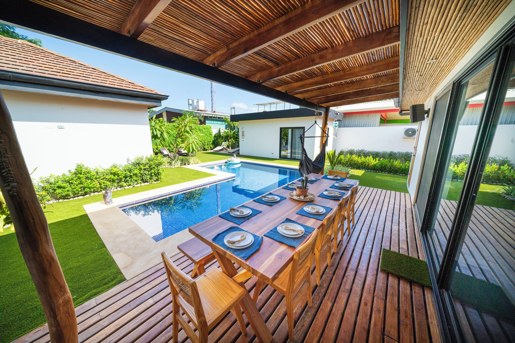 A wooden deck with a pool table set up for dining, surrounded by a pool and greenery.