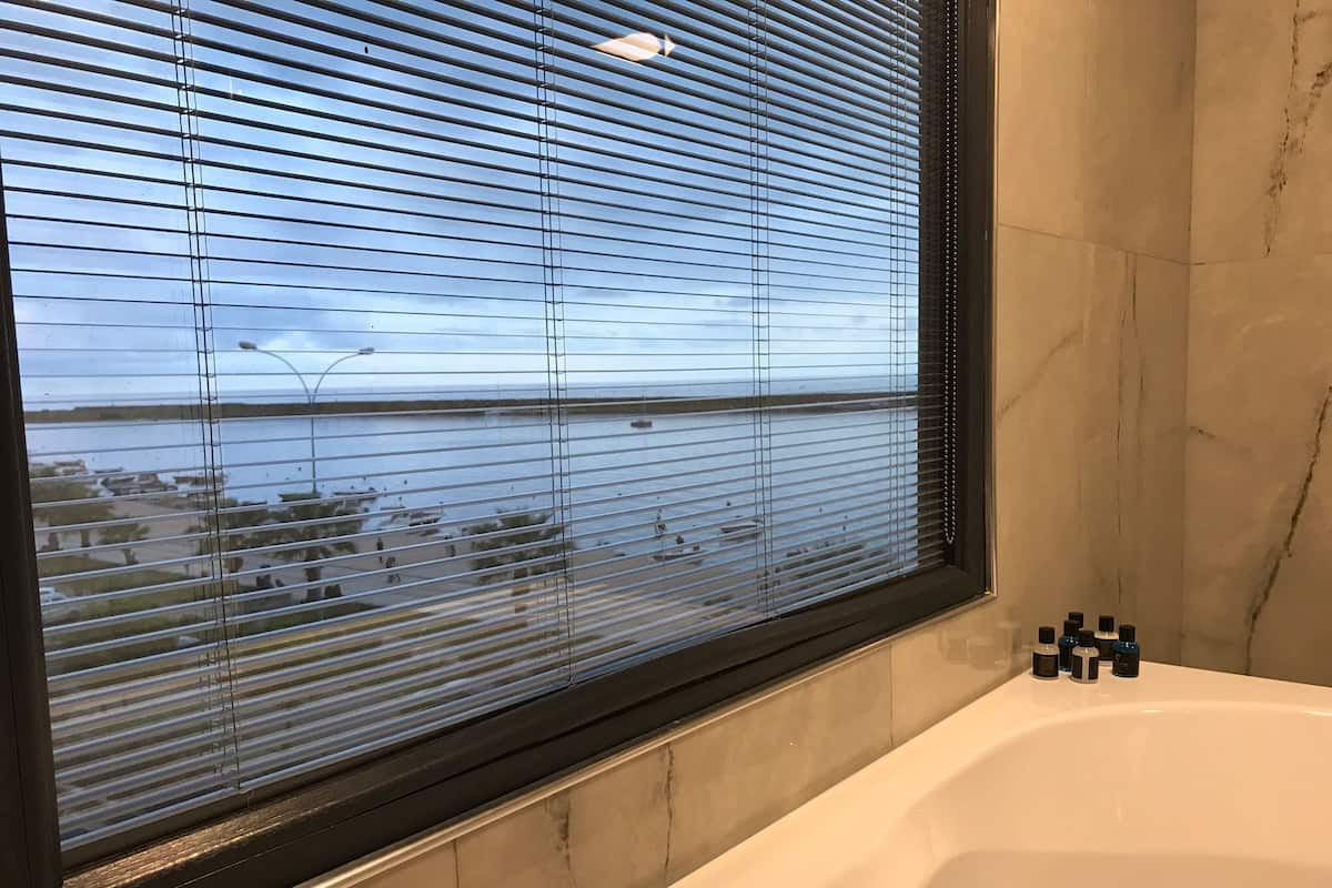 A bathroom with a bathtub, a window with blinds, and a view of a body of water.