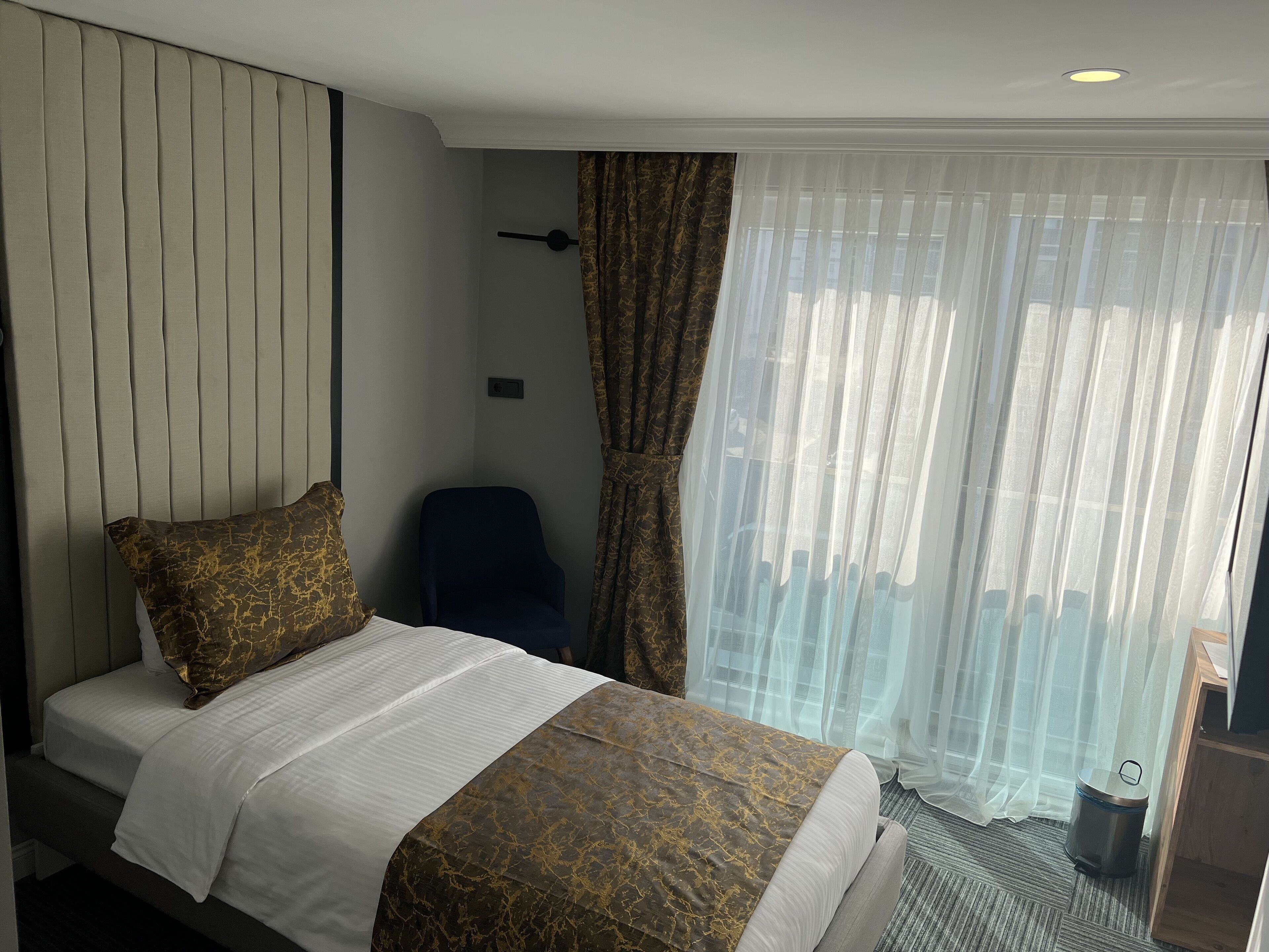 A hotel room with a bed, a chair, a window with sheer curtains, and a small trash bin.