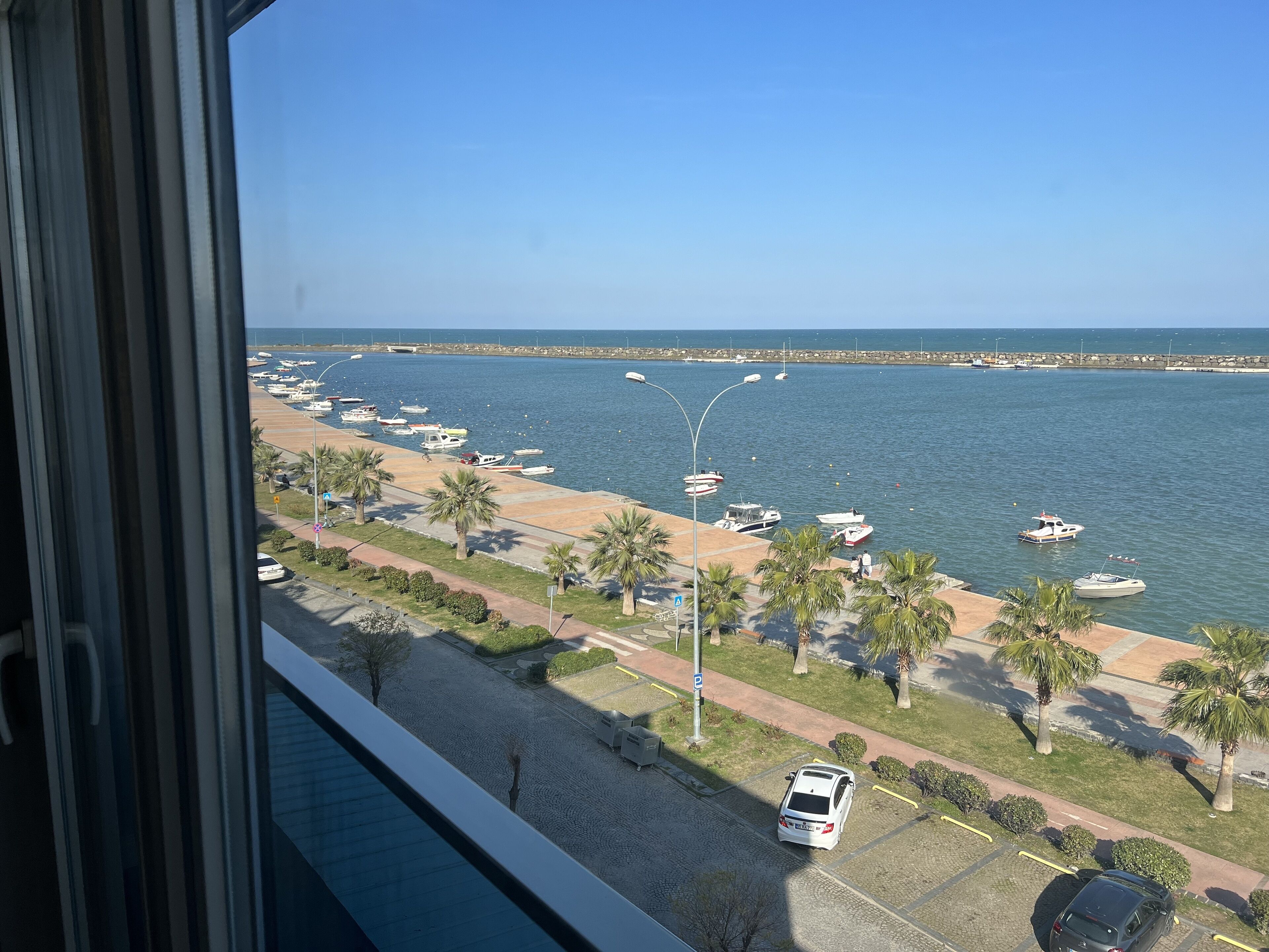 A coastal view with boats docked along the shore, a parking area with a few cars, and a clear sky.