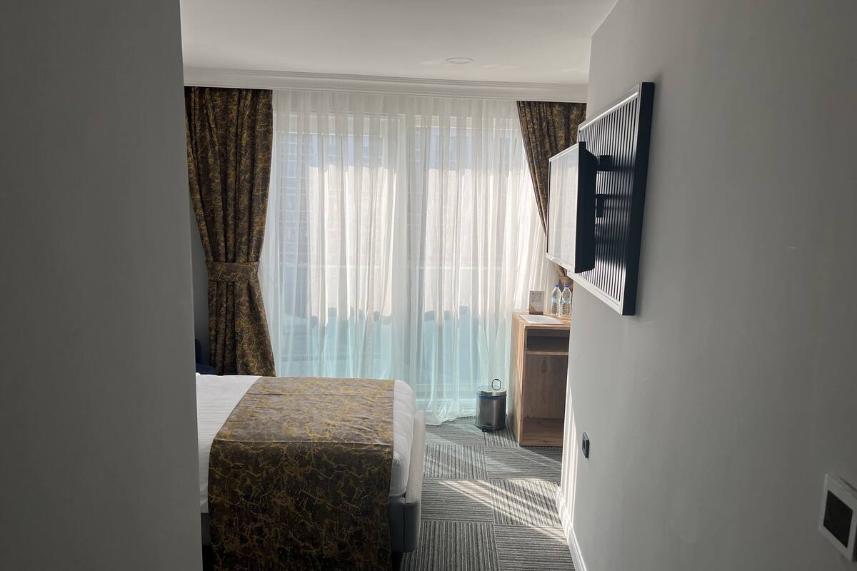 A hotel room with a bed, a small desk, a trash bin, and a window with sheer curtains.