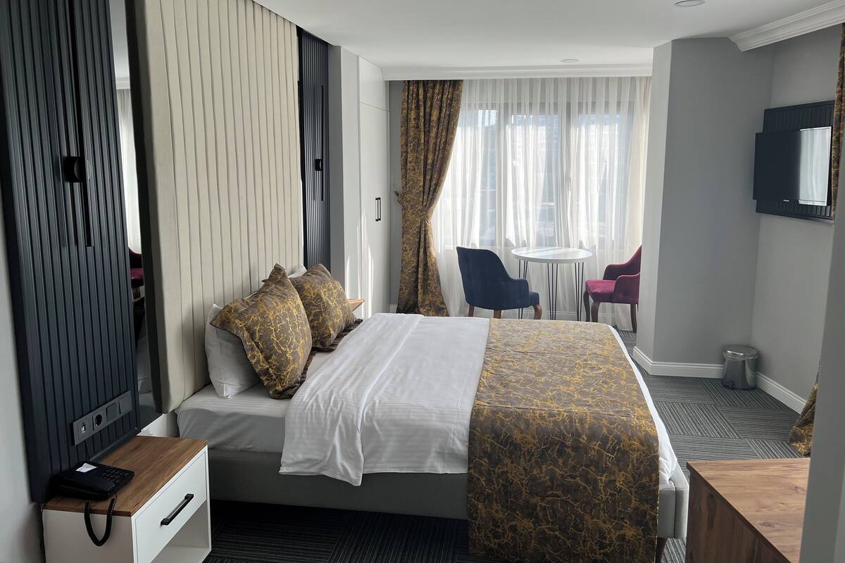 A hotel room with a bed, bedside table, and a desk with a chair. There is a window with curtains and a view of the outside.