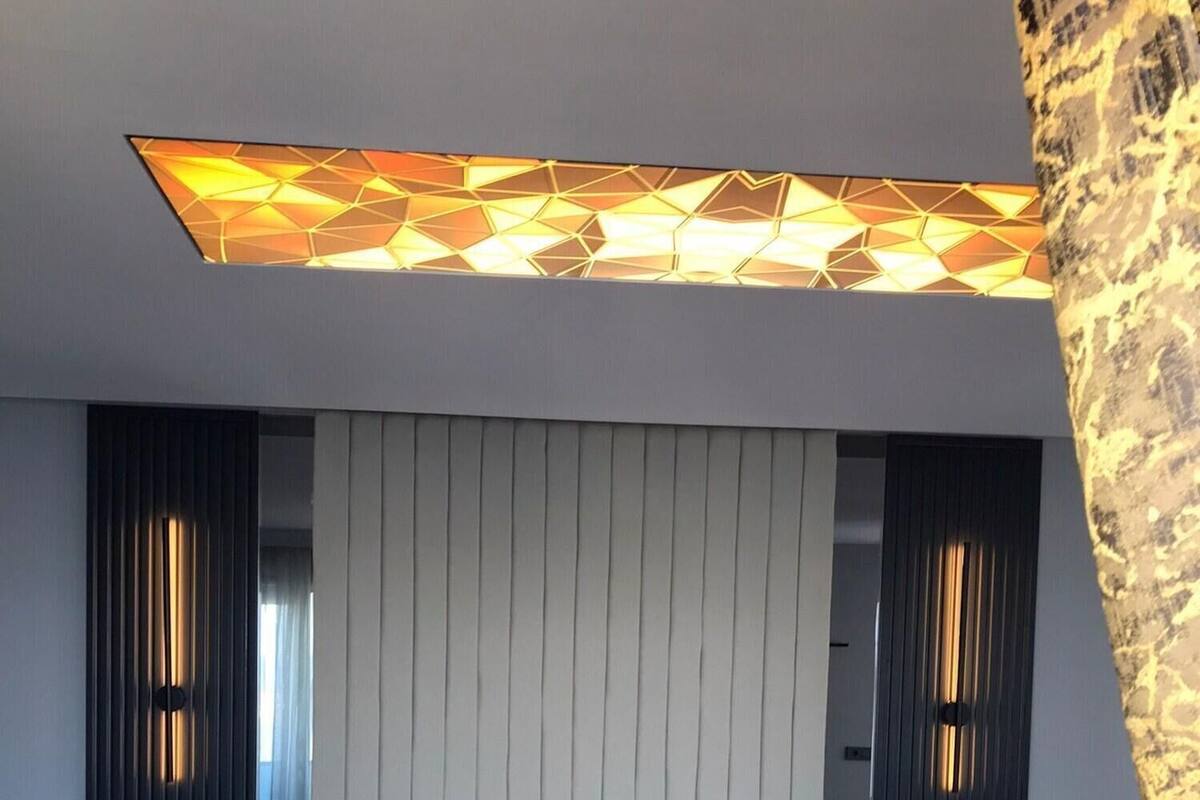 A modern hotel room with a large bed, two nightstands, a headboard with vertical lines, and a geometric-patterned ceiling light.