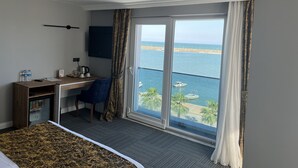 A hotel room with a bed, a desk with a chair, a television, a window with curtains, and a view of the sea. - Cepnis Hotel (Atakum)