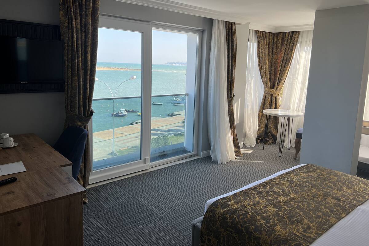 A hotel room with a large sliding glass door to a balcony overlooking the sea, a bed with a patterned bedspread, a wooden desk with a television, and a small table with a cup and saucer.