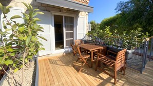 Una terraza de madera con una mesa y sillas, un edificio blanco con techo de tejas y follaje a su alrededor.