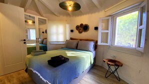 Un dormitorio con cama, una mesita auxiliar, una ventana con vistas, una lámpara en el techo y una puerta que da a otra habitación.