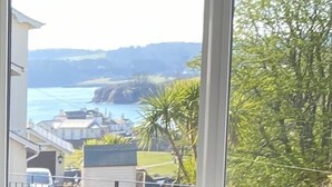 A room with a view of a coastal landscape through large windows, a black metal chair with cushions, and a framed picture on the windowsill. - Seaford Sands holiday home with sea view and on South West Coast path (Paignton)
