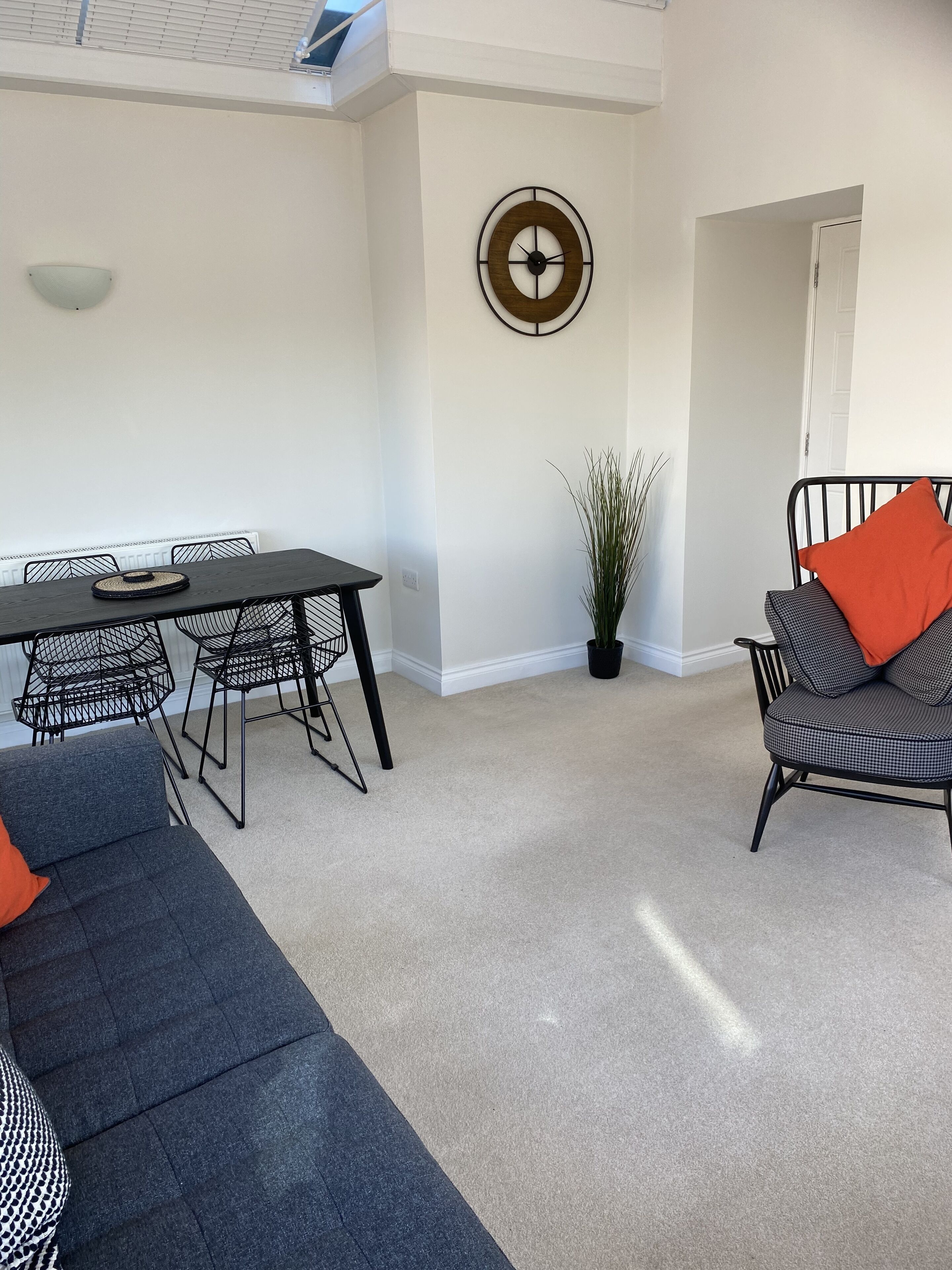 A modern living room with a dark grey sofa, a black dining table with metal chairs, a potted plant, and a wall clock.
