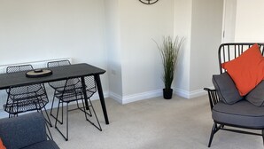 A modern living room with a dark grey sofa, a black dining table with metal chairs, a potted plant, and a wall clock. - Seaford Sands holiday home with sea view and on South West Coast path (Paignton)
