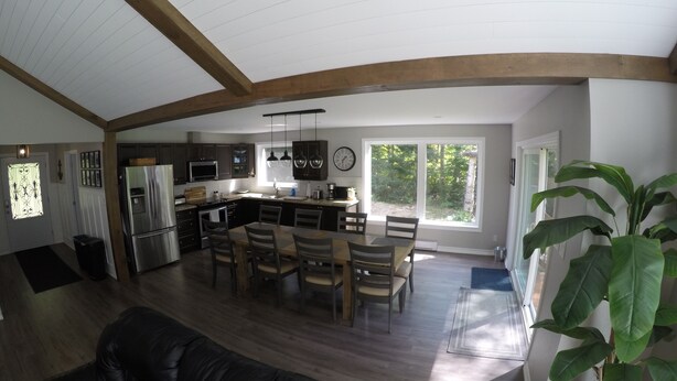 A spacious living area with a dining table and chairs, a kitchen with modern appliances, and large windows offering a view of the outdoors. - MODERN COTTAGE GETAWAY (Barry's Bay)