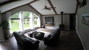 A spacious living room with a high ceiling, a fireplace, and a large window overlooking a garden. - MODERN COTTAGE GETAWAY (Barry's Bay)