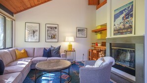 A cozy living room with a sofa, armchair, and a round coffee table. There are framed pictures on the wall, a lamp, and a fireplace.