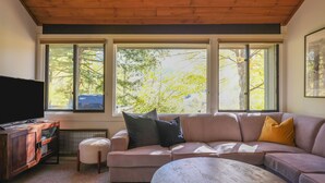 A living room with a large sectional sofa, a coffee table, a TV, and a fireplace. There are large windows with a view of trees and a blue sky.