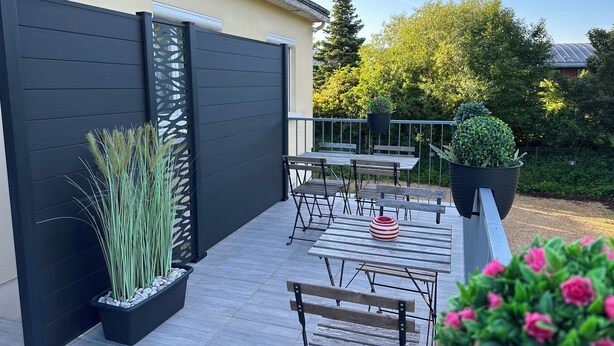 Eine Terrasse mit Holztischen und -bänken, umgeben von einem schwarzen Zaun und Kübelpflanzen.