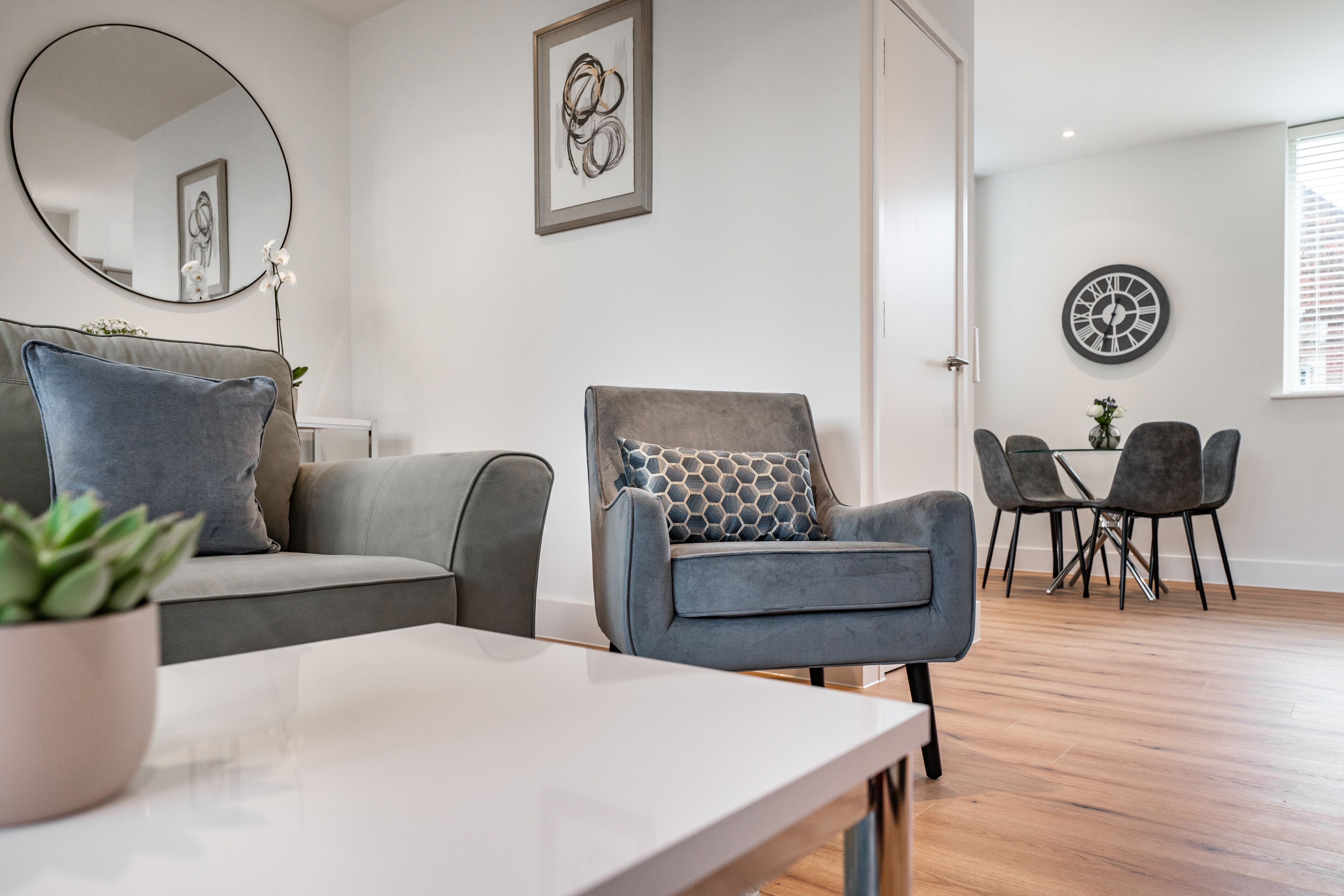 A modern living room with a grey sofa, a grey armchair, a white coffee table, and a dining area with a glass table and grey chairs.