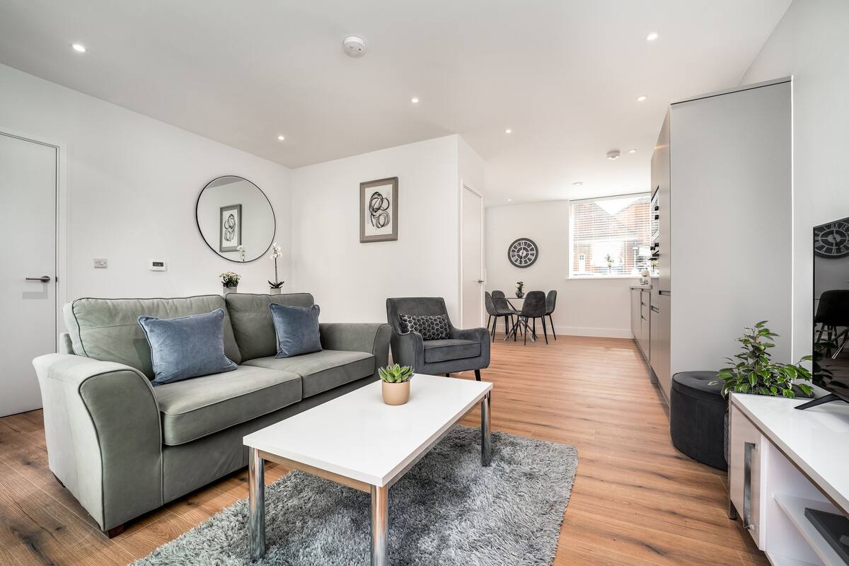 A modern living room with a grey sofa, a white coffee table, and a black armchair. There is a dining area with a table and chairs in the background.