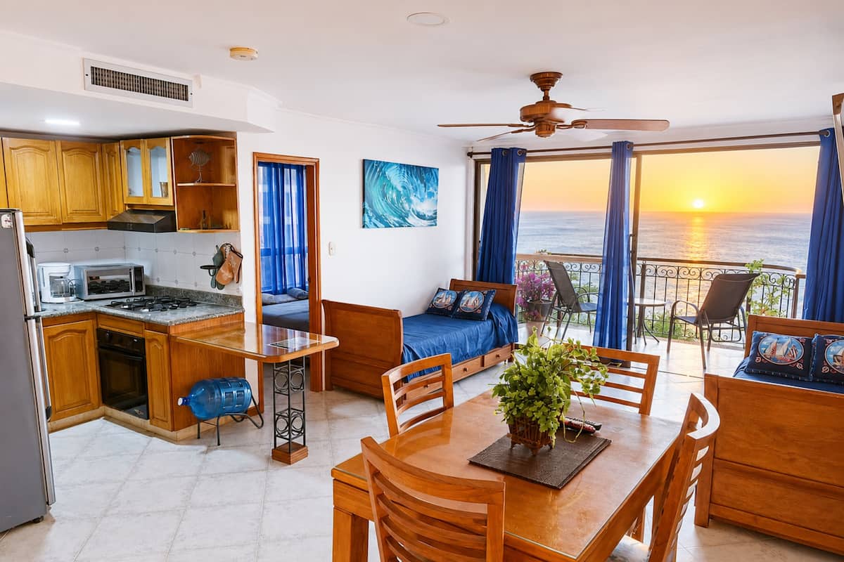 Un espace de vie compact comprenant un coin cuisine, un coin repas, une chambre avec vue sur la mer et un balcon aménagé avec des sièges.