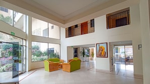 Un hall d’entrée spacieux, doté de hauts plafonds, de grandes fenêtres et offrant une vue sur l’extérieur. On y trouve des fauteuils en osier et une table au centre, ainsi qu’un tableau accroché au mur.