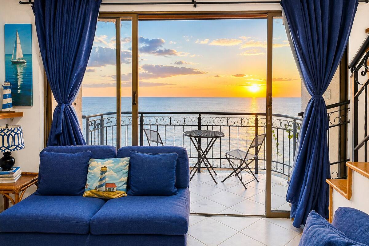 Un balcon avec vue sur le coucher de soleil sur la mer, un canapé bleu avec des coussins et une petite table avec des chaises.