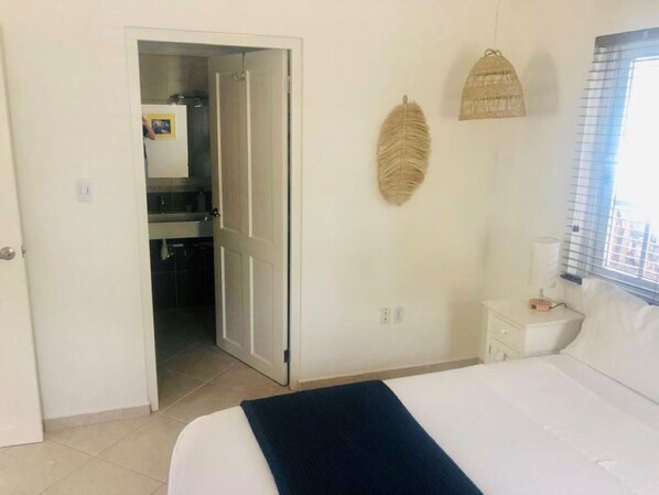 A bedroom with a bed, a nightstand, a wall-mounted lamp, a door leading to a bathroom, and a woven wall decoration.
