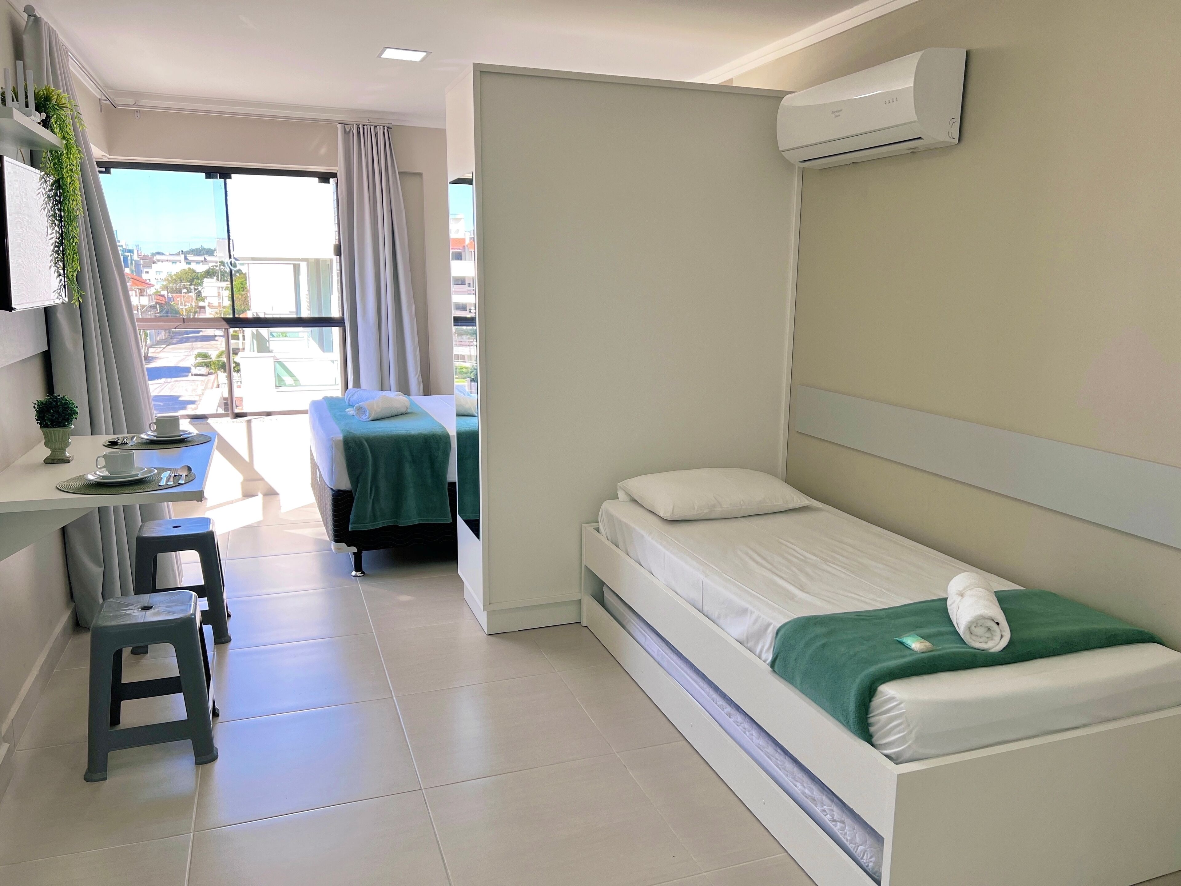 Une chambre d’hôtel comprenant un lit, un bureau avec une chaise, un petit tabouret, un appareil de climatisation et une vue sur la ville par la fenêtre.