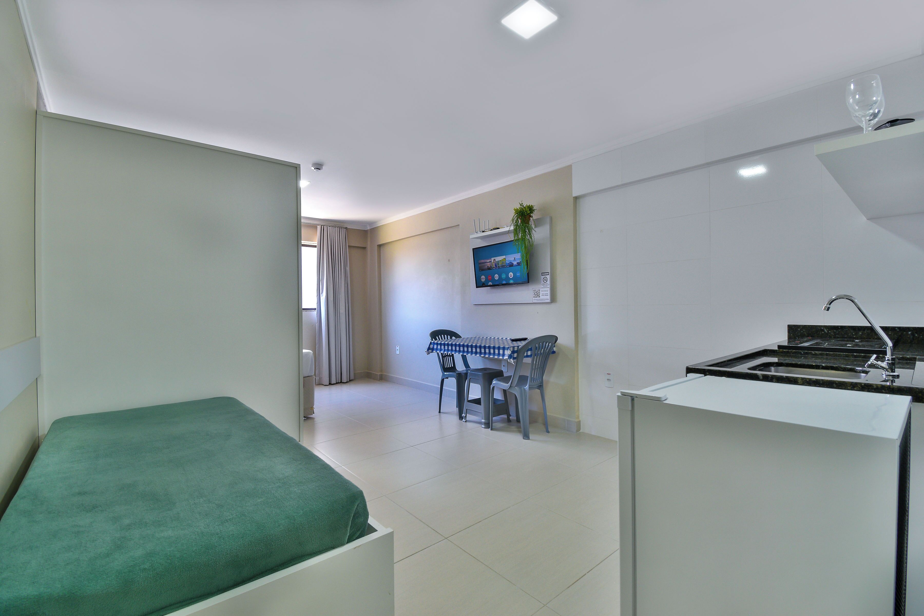 A compact hotel room with a green bed, a small kitchenette, a dining area with a table and chairs, and a TV mounted on the wall.