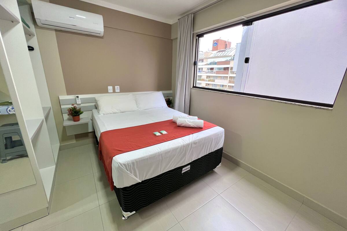 A hotel room with a bed, a window, a white air conditioner, and a small table with a plant.