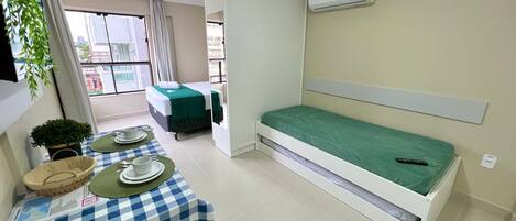 A compact hotel room with a single bed, a small table with a checkered tablecloth, a potted plant, and a window with curtains.