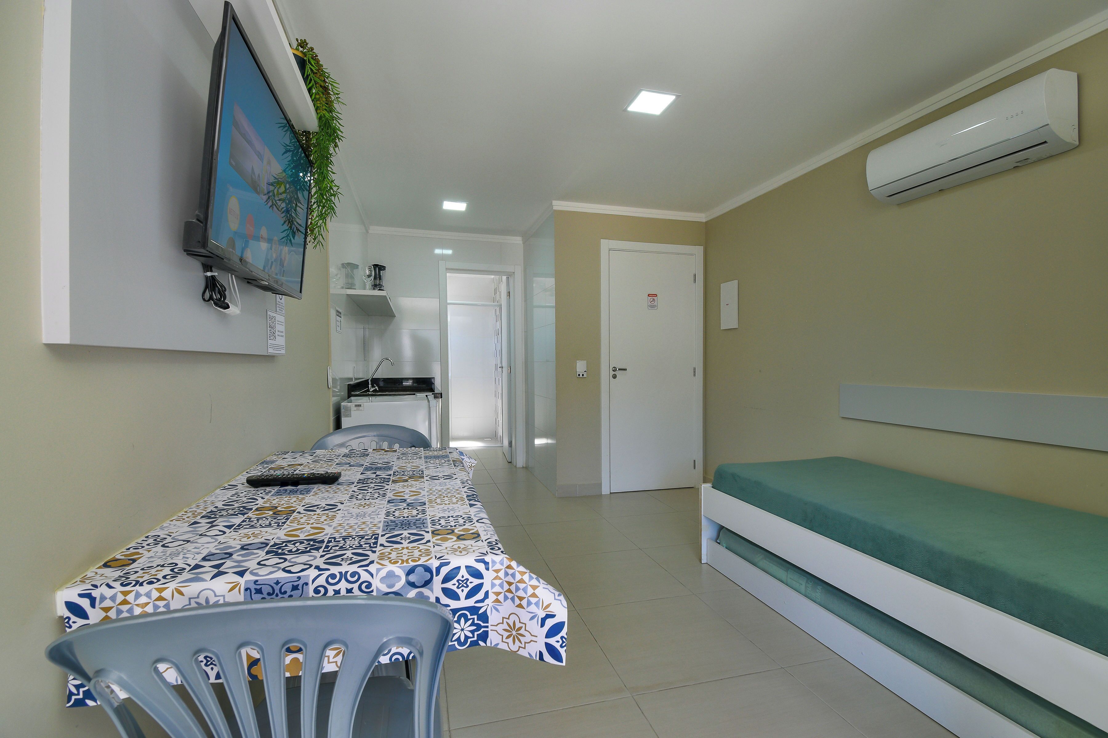 A hotel room with a bed, a table with a patterned tablecloth, a chair, an air conditioning unit, a television mounted on the wall, and a door leading to another room.