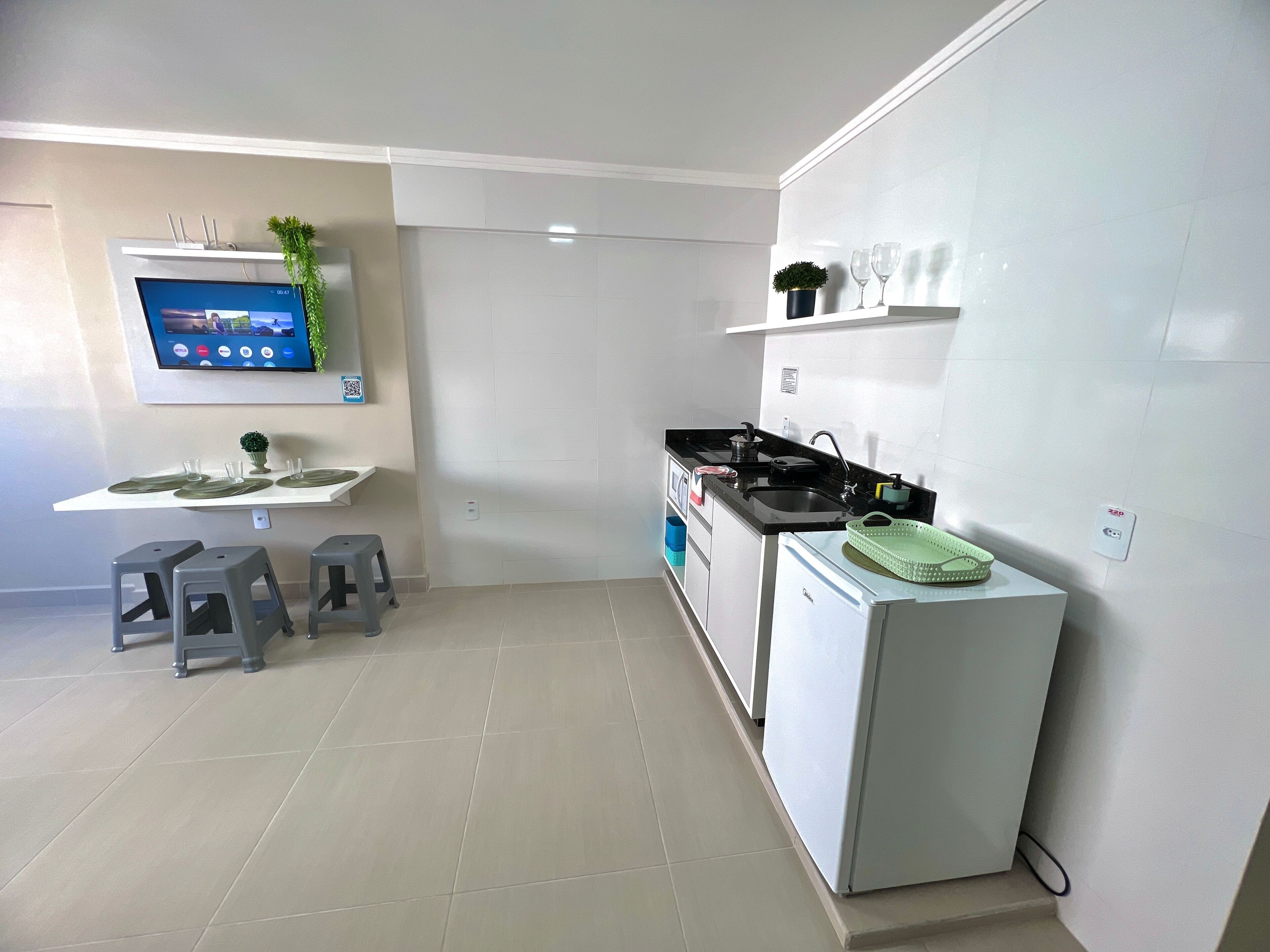 Cocina moderna con horno empotrado, lavamanos y un refrigerador pequeño. Se observa un televisor de pantalla plana montado en la pared, una planta en maceta y un par de taburetes.