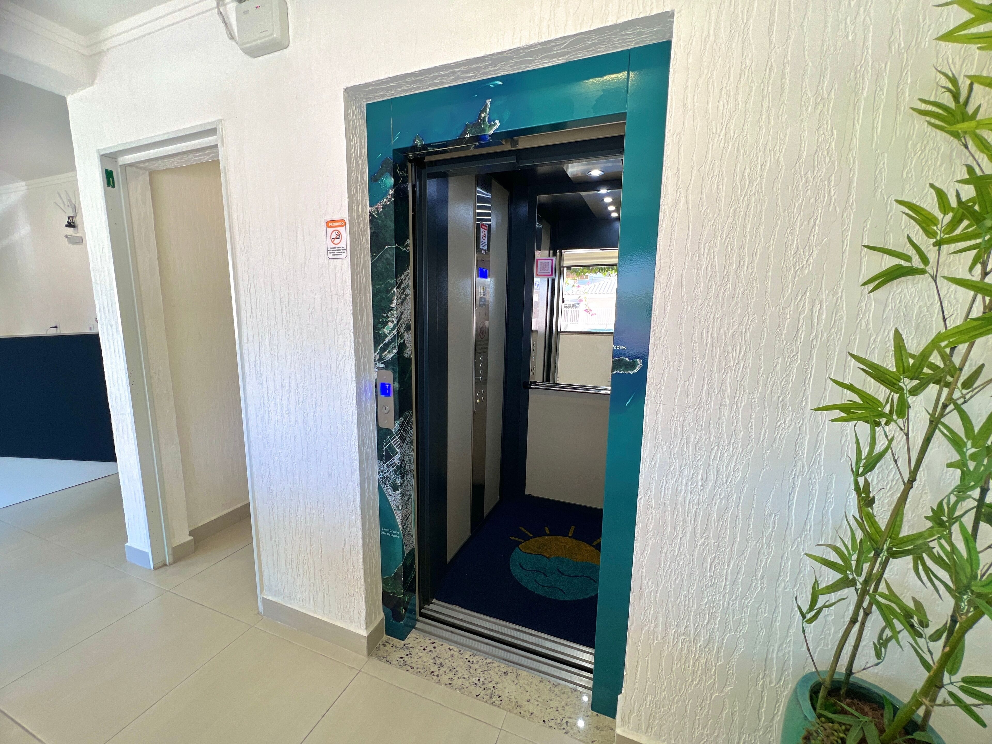 Ascensor de hotel con una puerta color turquesa, un letrero de salida de emergencia visible y una planta en maceta a su lado.