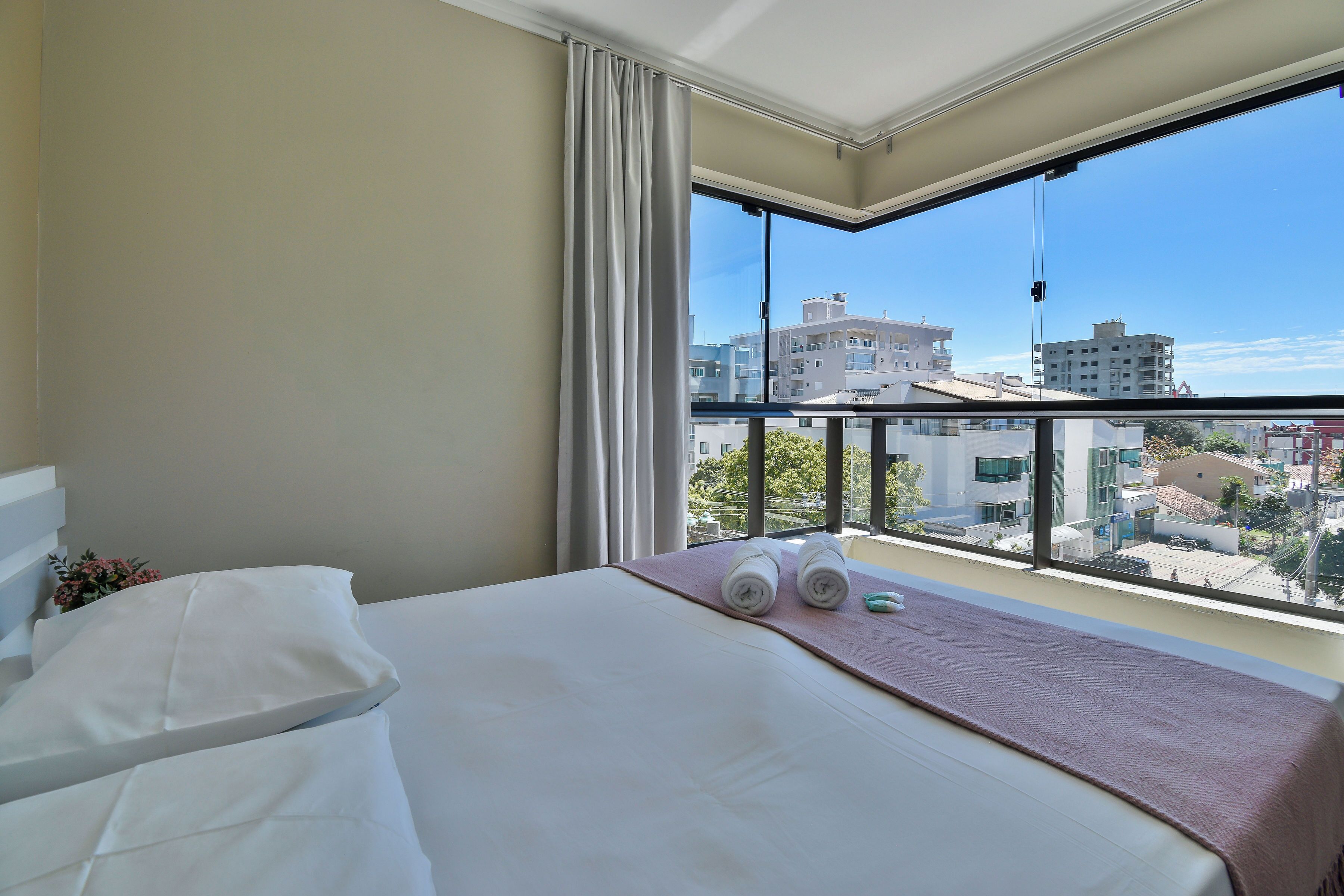 Une chambre d’hôtel avec une grande fenêtre, un lit avec du linge de maison blanc, une serviette rose et une vue sur les immeubles.