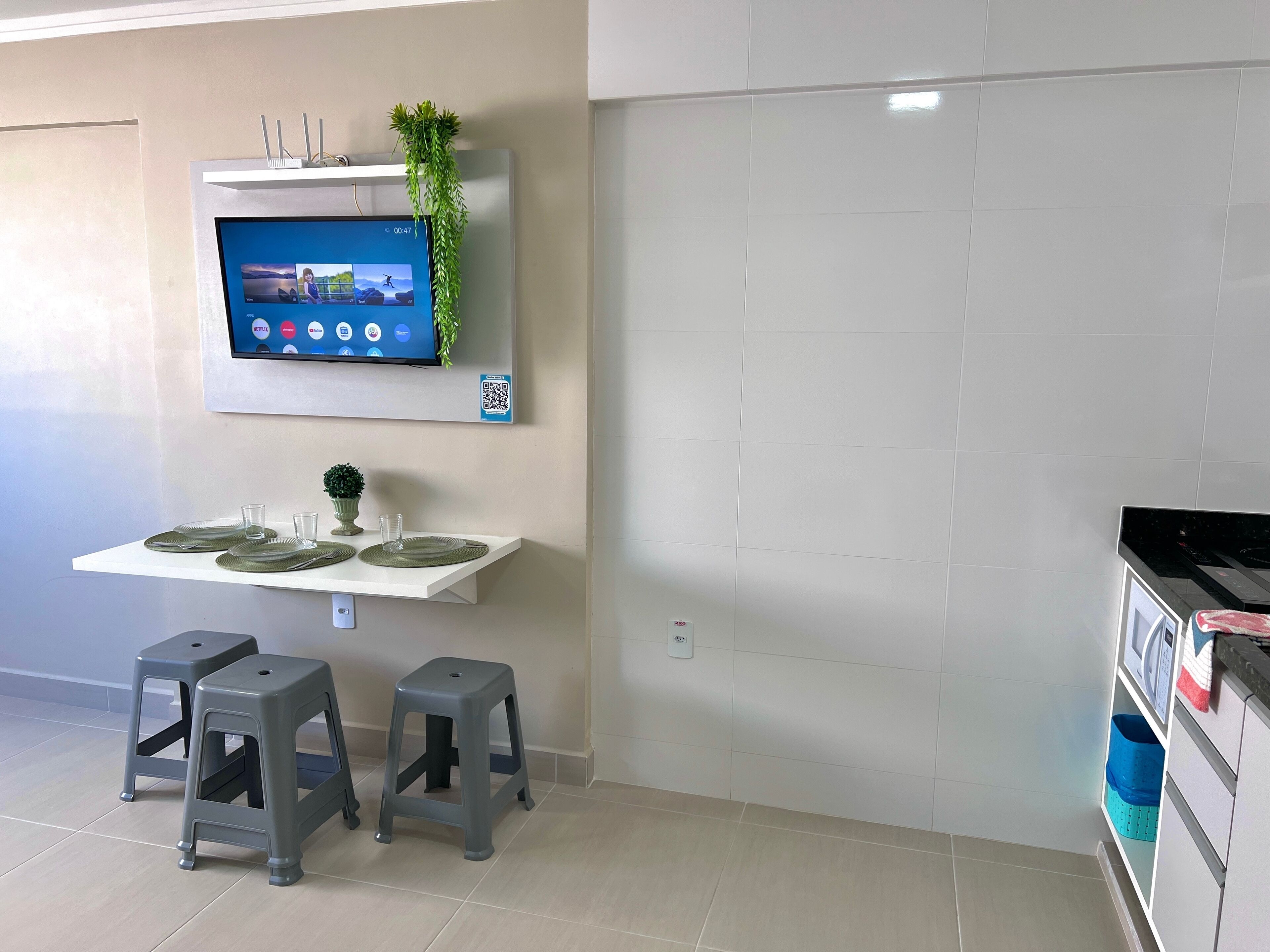 Una cocina moderna con un televisor de pantalla plana montado en la pared, una planta pequeña y un código QR. Hay dos taburetes y una mesa con platos y vasos.