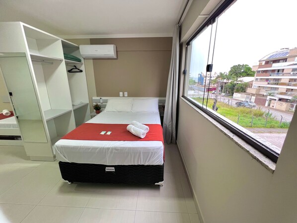 A hotel room with a bed, a wardrobe, a window with a view of buildings, and an air conditioning unit. - Canto da Catarina Bombinhas (Bombinhas)