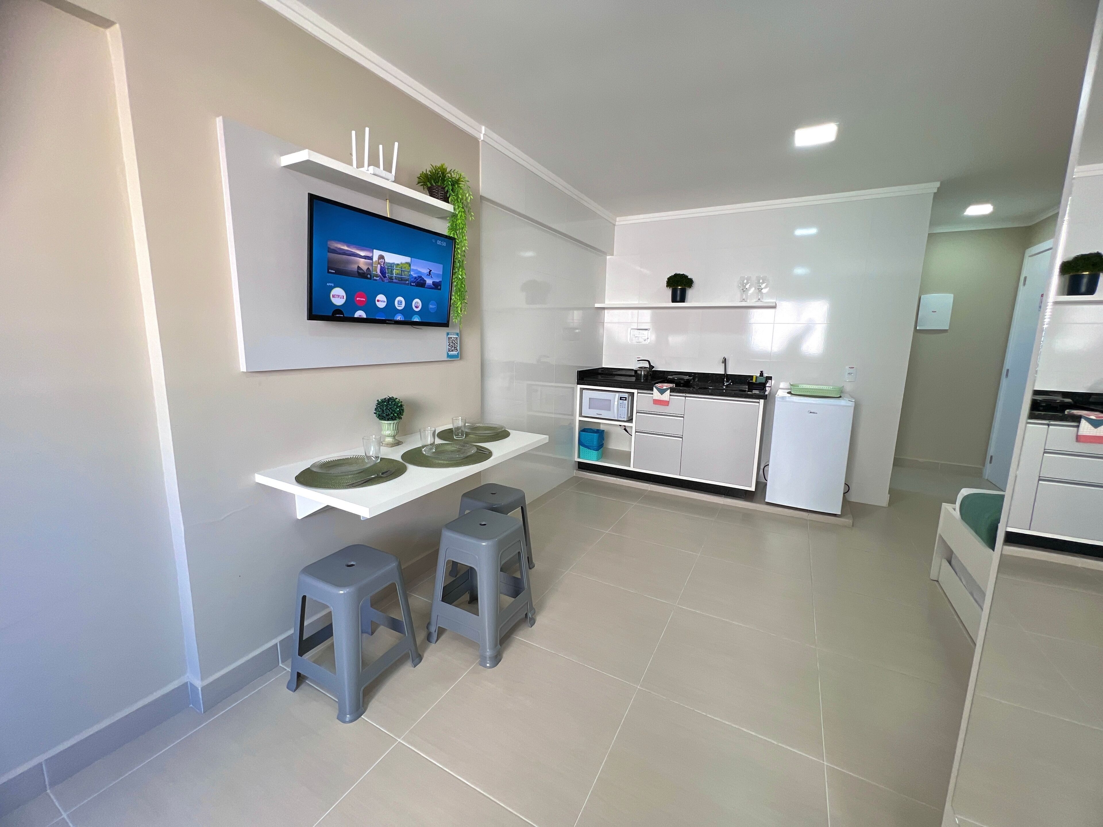 Una cocina moderna con televisor integrado, un pequeño comedor con taburetes y un refrigerador compacto.