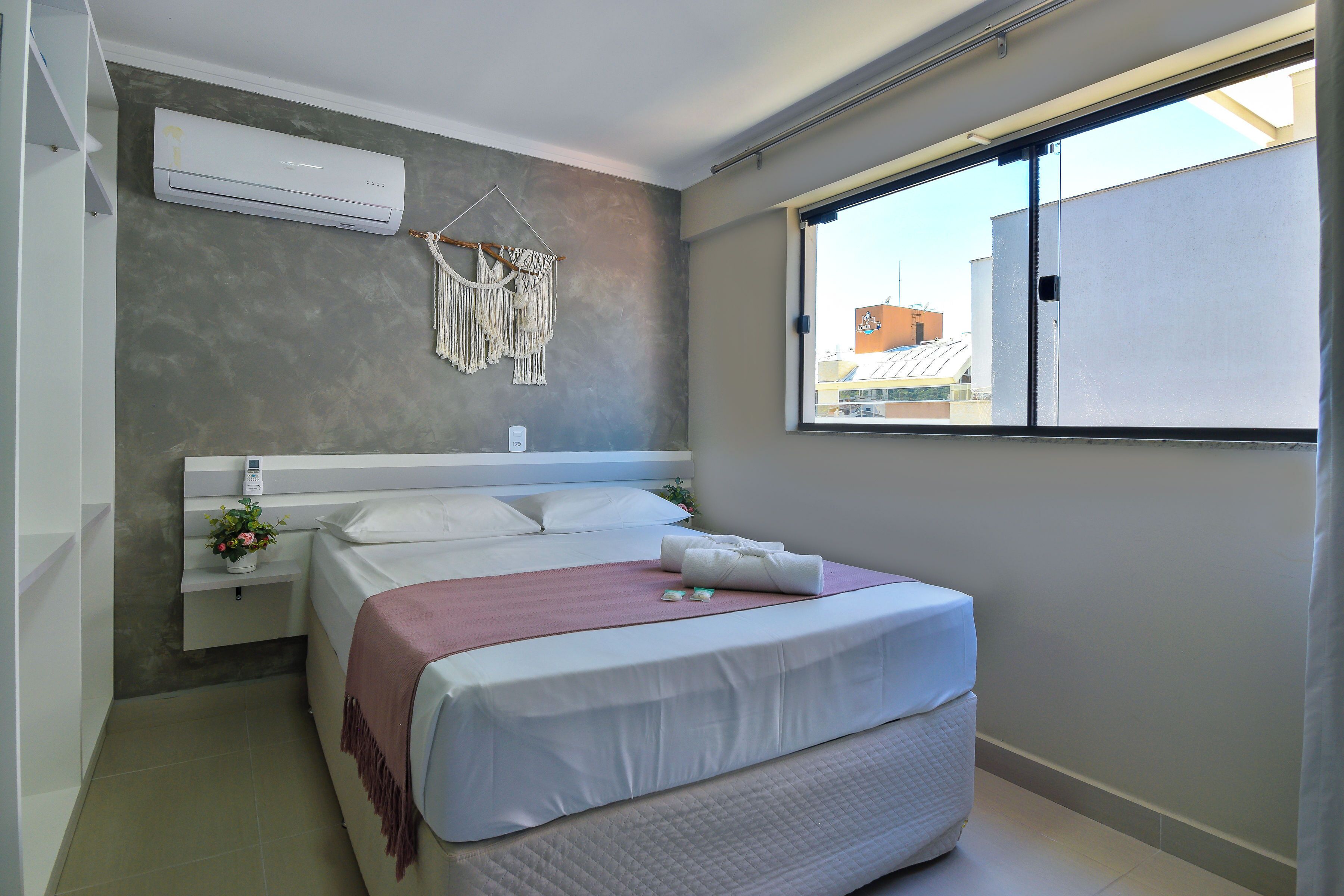 A bedroom with a bed, bedside table, air conditioning unit, macrame wall hanging, and a window with a view of buildings.