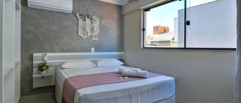 A bedroom with a bed, bedside table, air conditioning unit, macrame wall hanging, and a window with a view of buildings.
