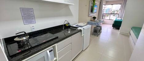 Una cocina moderna con encimera negra, armarios blancos, microondas y un refrigerador pequeño. Hay una planta en maceta sobre la encimera y una ventana con vistas al exterior.
